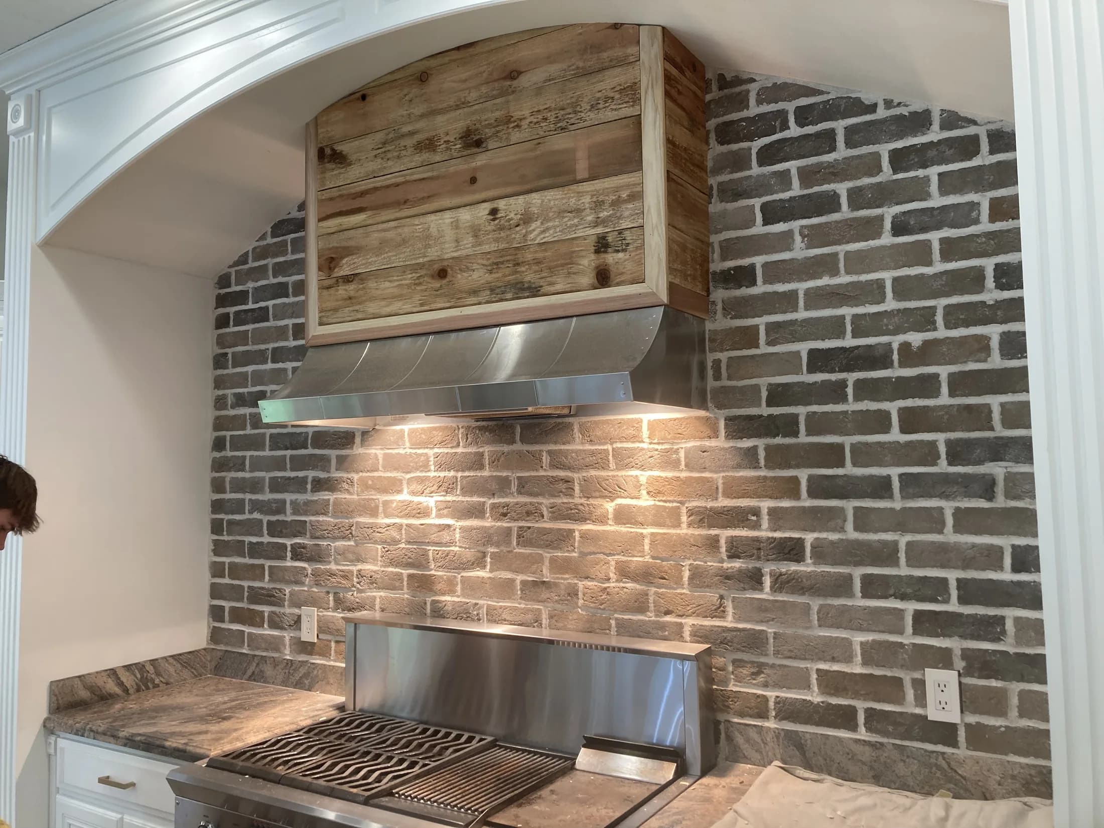 Custom wood range hood centered on a hand-finished gray-brick kitchen wall